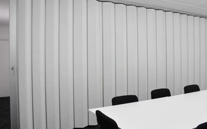 A white accordion door drawn closed across a meeting room, hanging from a single top track with no floor channel