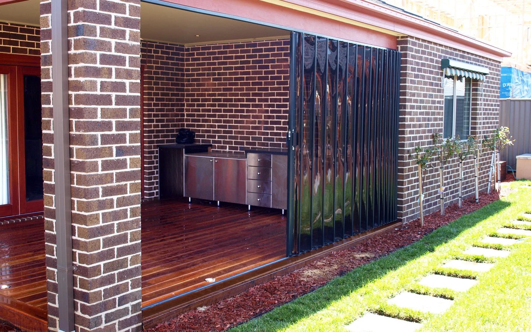 Clear folding outdoor blinds closed across the open side of a brick alfresco area, seen from the garden